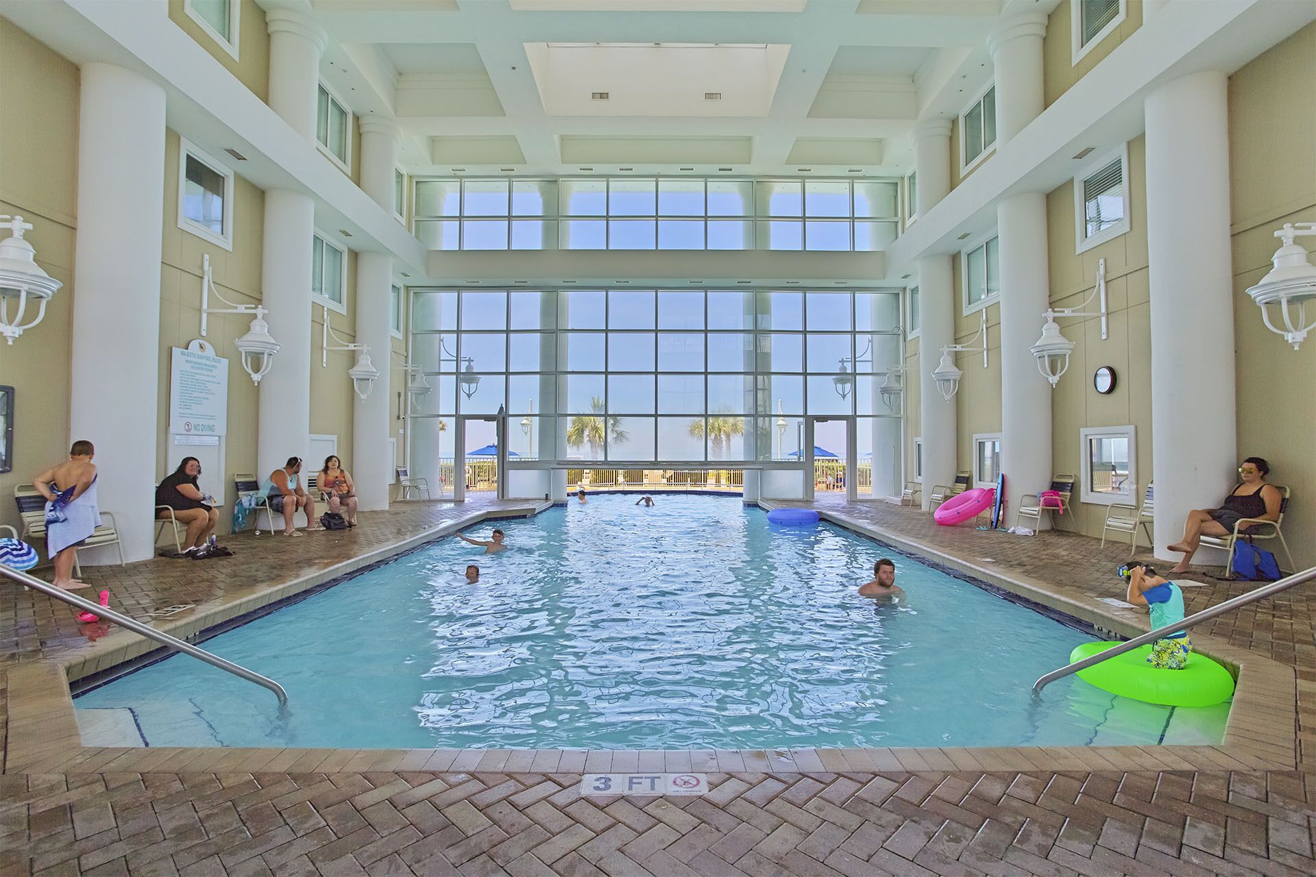 An indoor swimming pool with clear glass walls, a tiled floor, and a few people swimming and relaxing.
