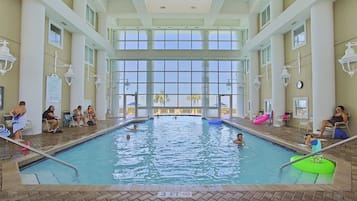 An indoor swimming pool with clear glass walls, a tiled floor, and a few people swimming and relaxing.