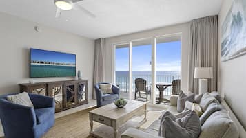 A spacious living room with a large TV, comfortable seating, a wooden coffee table, and a view of the ocean through large windows.