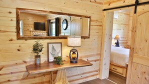 Un interior de madera con un espejo, una mesa con lámpara, un cuadro enmarco y una planta pequeña.