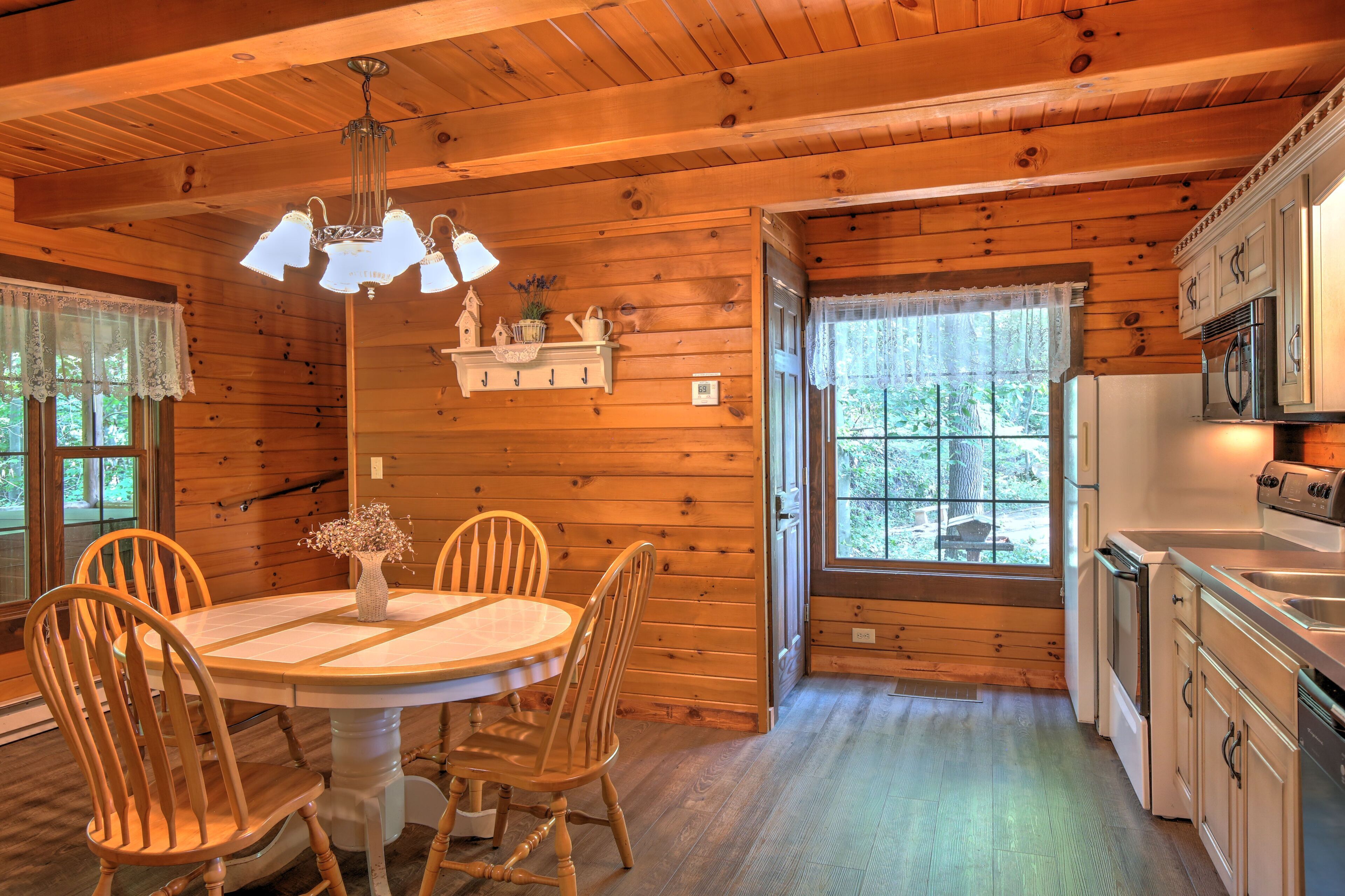 A wooden cabin interior with a dining table, chairs, a chandelier, and a kitchen area.