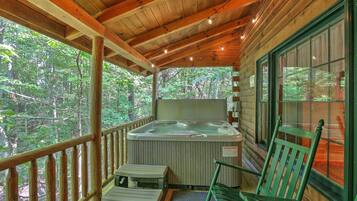 A wooden deck with a hot tub, wooden chairs, and a view of the forest.