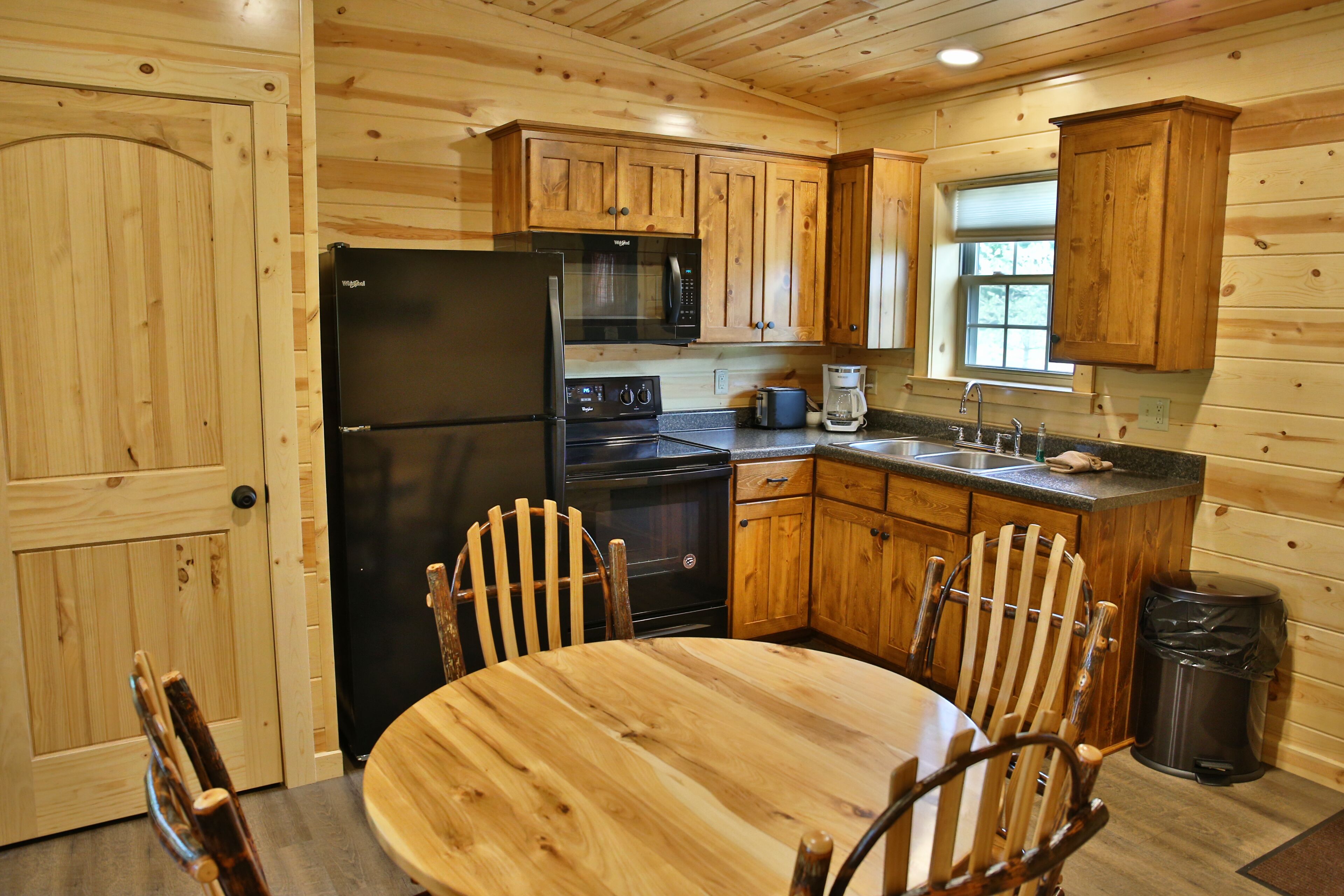 Cocina de cabaña de madera con refrigerador negro, microondas y estufa, armarios de madera, una mesa de comedor con cuatro sillas y una ventana.