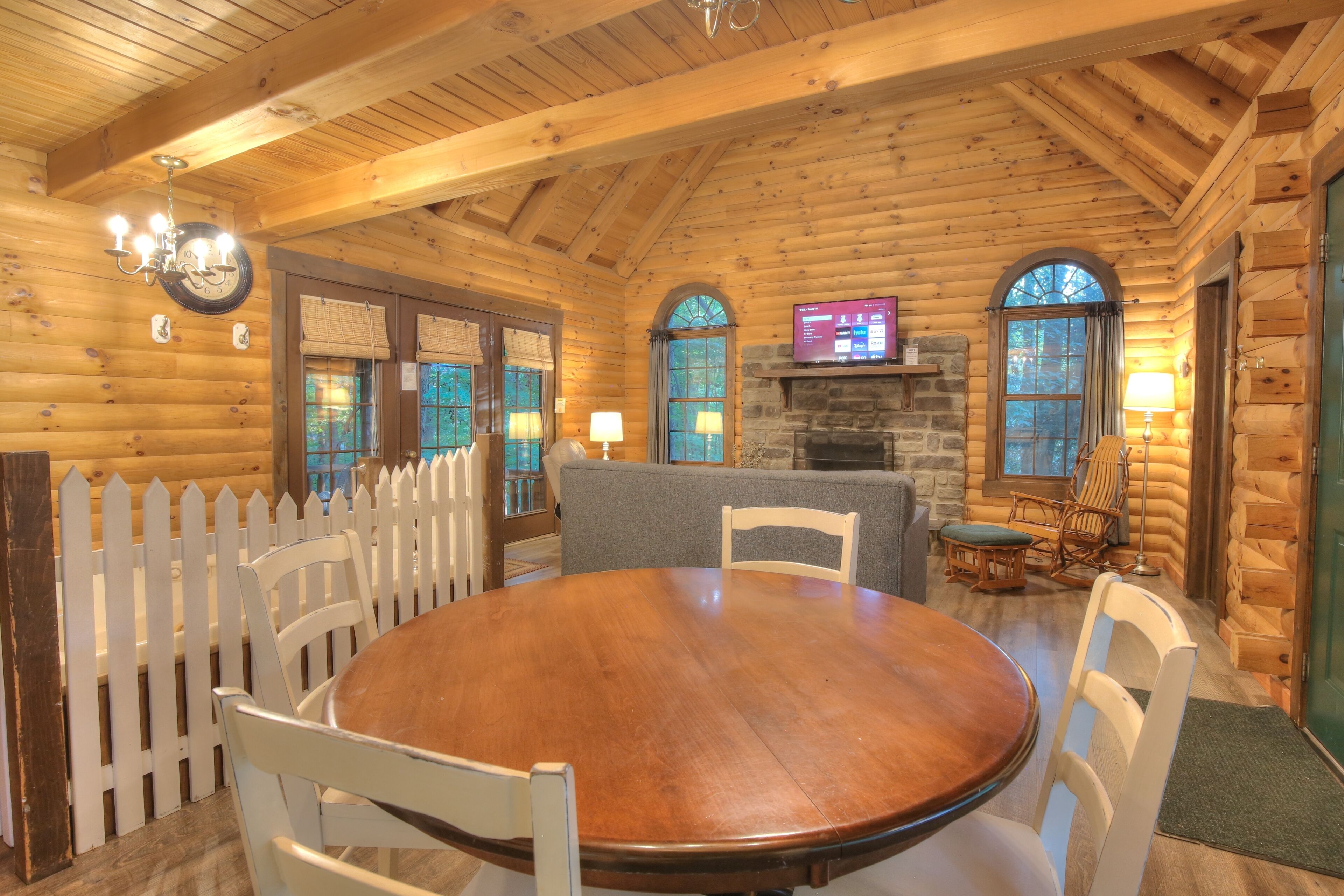 A wooden cabin interior with a round wooden table, white chairs, a stone fireplace, and a television mounted on the wall.