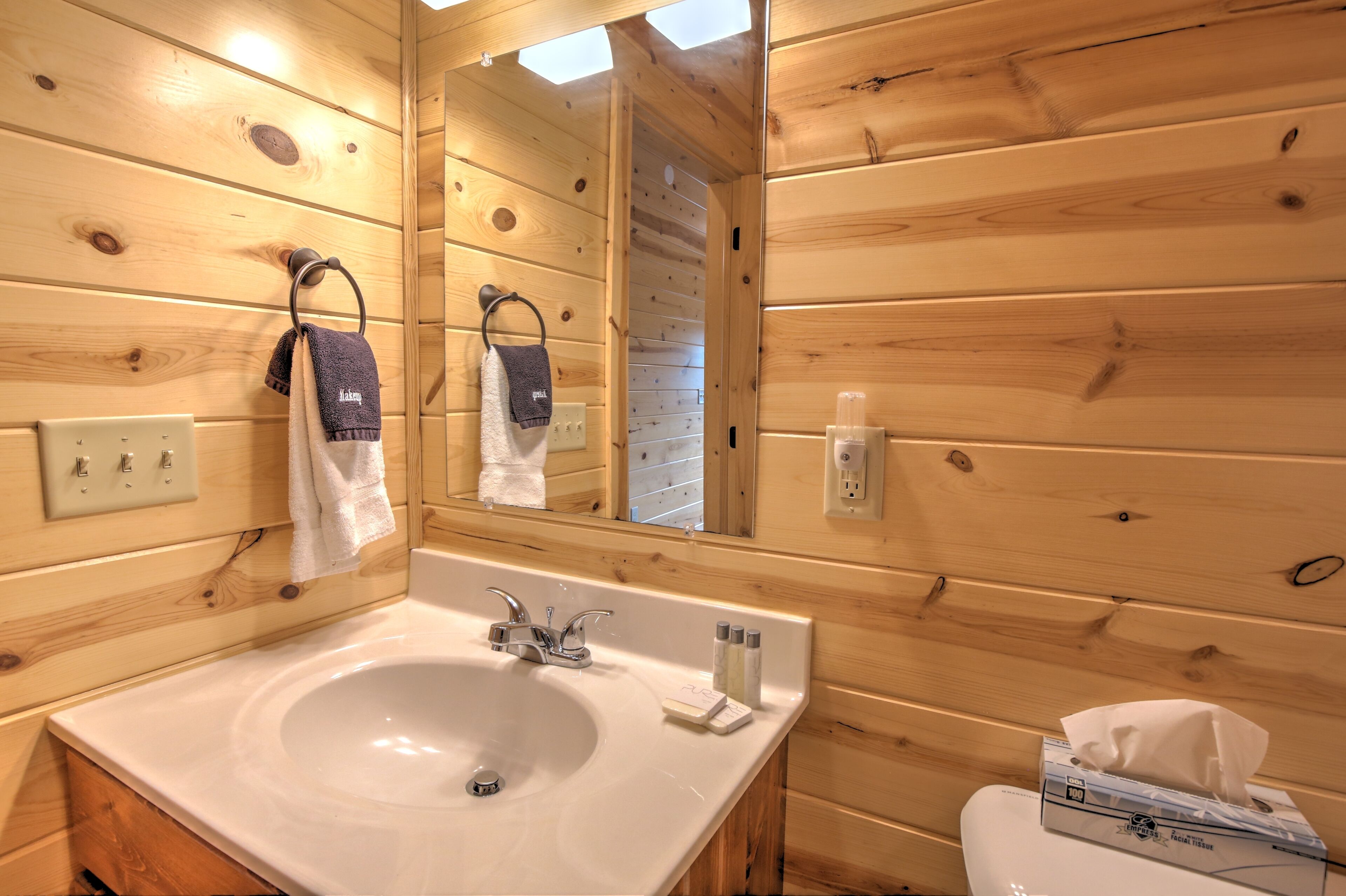 Ein Badezimmer einer Holzhütte mit Waschbecken, Spiegel, Handtuchringen und einer Taschentuchbox.