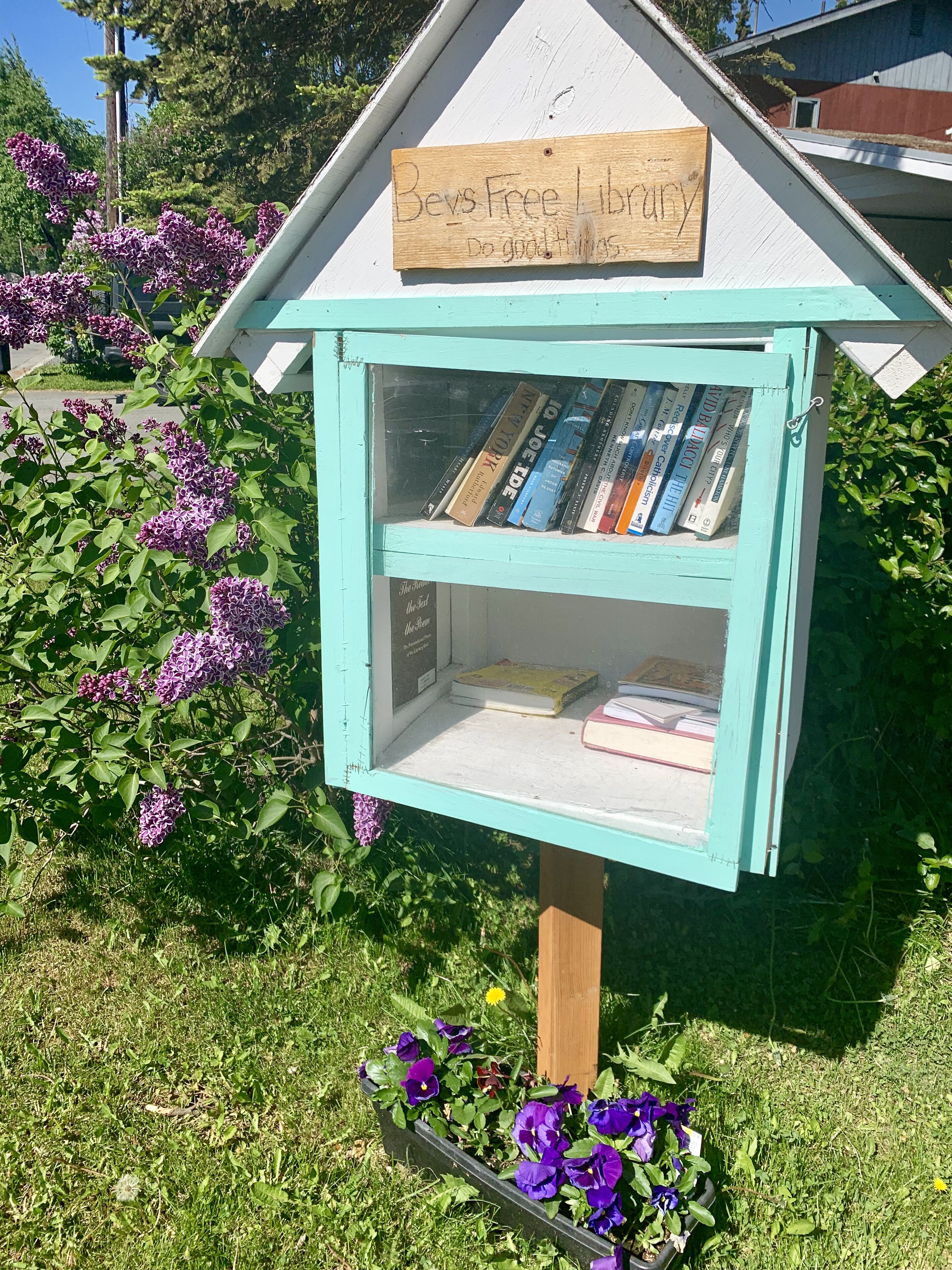A small, light blue library with a wooden sign that reads "Bevs Free Library" and a message "Do good things."