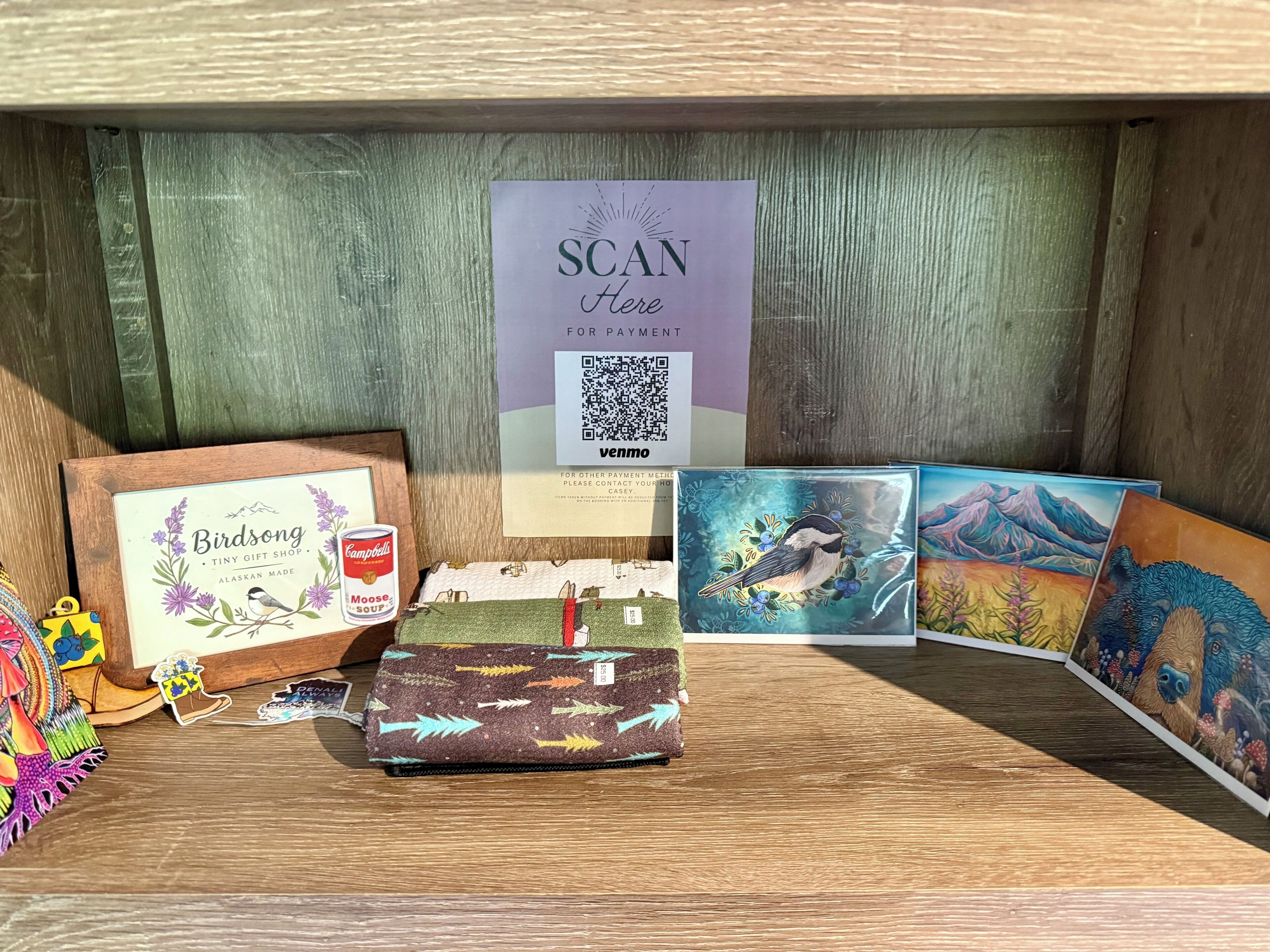 A display of various items including a framed Birdsong Tiny Gift Shop sign, a can of Campbell's Moose Soup, a wallet with arrows, and several cards with bird illustrations.