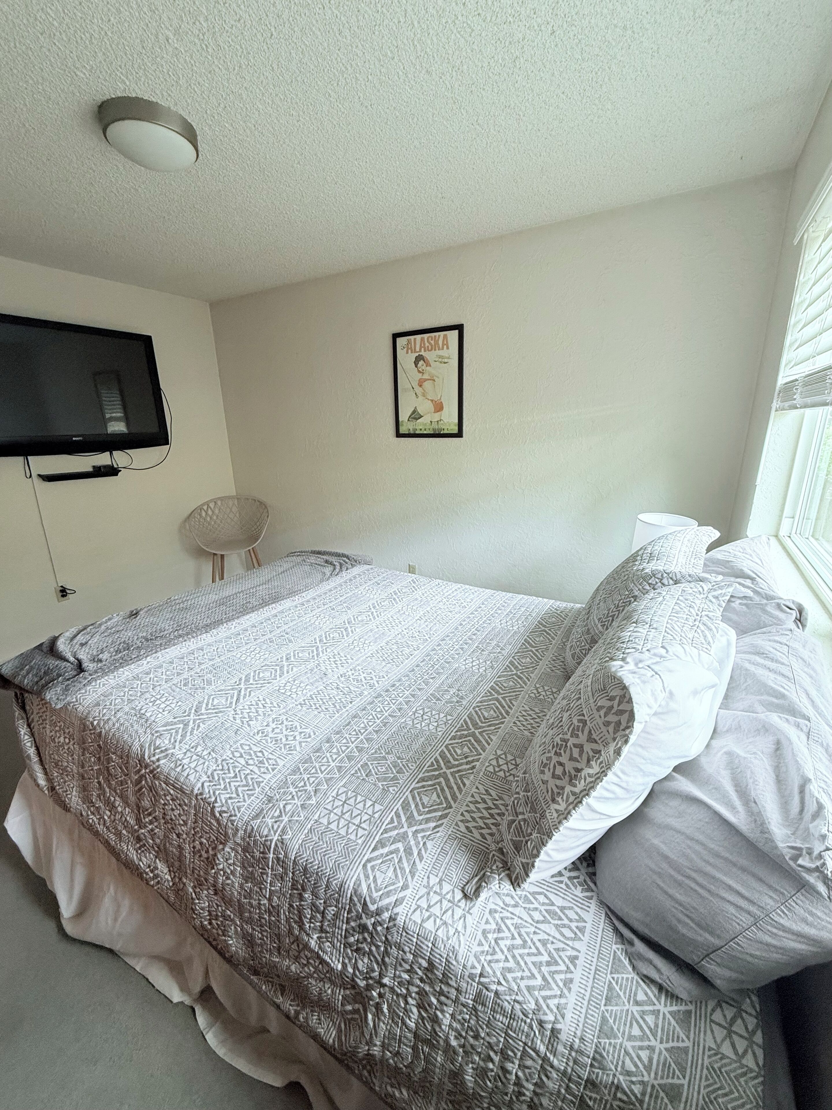Une chambre à coucher avec un lit, un téléviseur fixé au mur, un tableau encadré et une fenêtre avec des stores.
