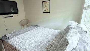 Une chambre à coucher avec un lit, un téléviseur fixé au mur, un tableau encadré et une fenêtre avec des stores.