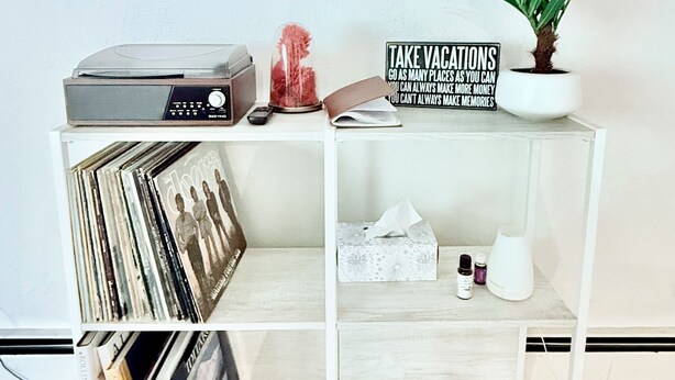Une étagère blanche sur laquelle se trouvent une platine vinyle, un cadre avec une citation, une plante en pot et un panneau rappelant l’importance de prendre des vacances.