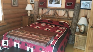 A log cabin-style bedroom with a bed, two bedside tables, a lamp, and wall decorations including framed pictures and a bear silhouette pillow.