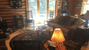 A cozy log cabin interior with a fireplace, wooden furniture, and a large window offering a view of trees.