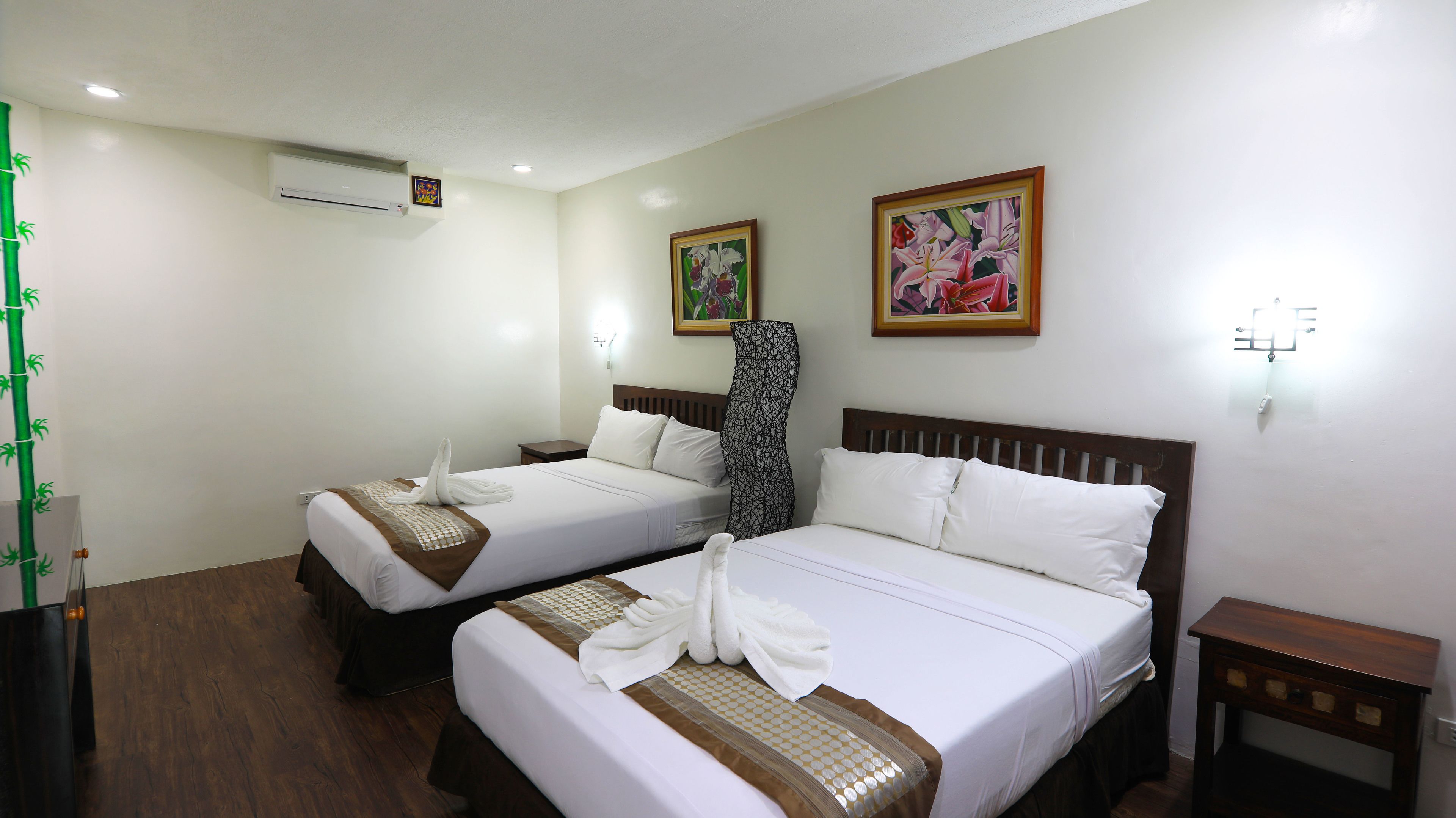 A hotel room with two beds, each with a neatly folded towel and a patterned bedspread. There are two paintings on the wall, a small table, and a wall-mounted air conditioner.