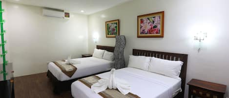 A hotel room with two beds, each with a neatly folded towel and a patterned bedspread. There are two paintings on the wall, a small table, and a wall-mounted air conditioner.