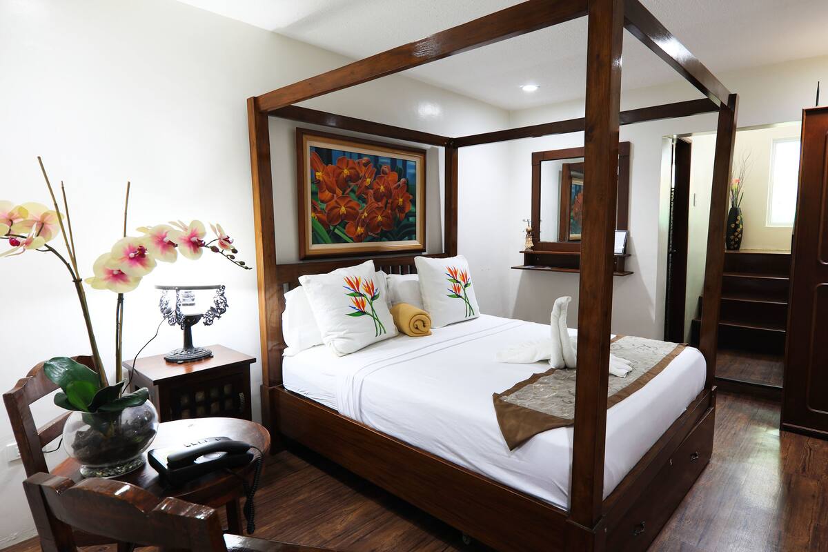 A four-poster bed with white linens, a painting above it, and a wooden wardrobe to the right.