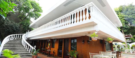 Un edificio di due piani con un balcone bianco, pareti in mattoni e un’area esterna con tavoli e sedie.