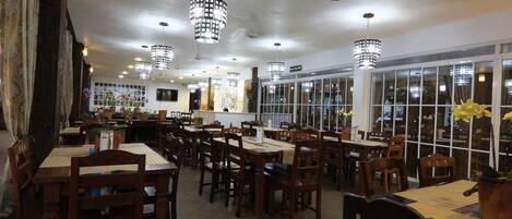 Een restaurant met meerdere houten tafels en stoelen, grote ramen en hangende lampen.