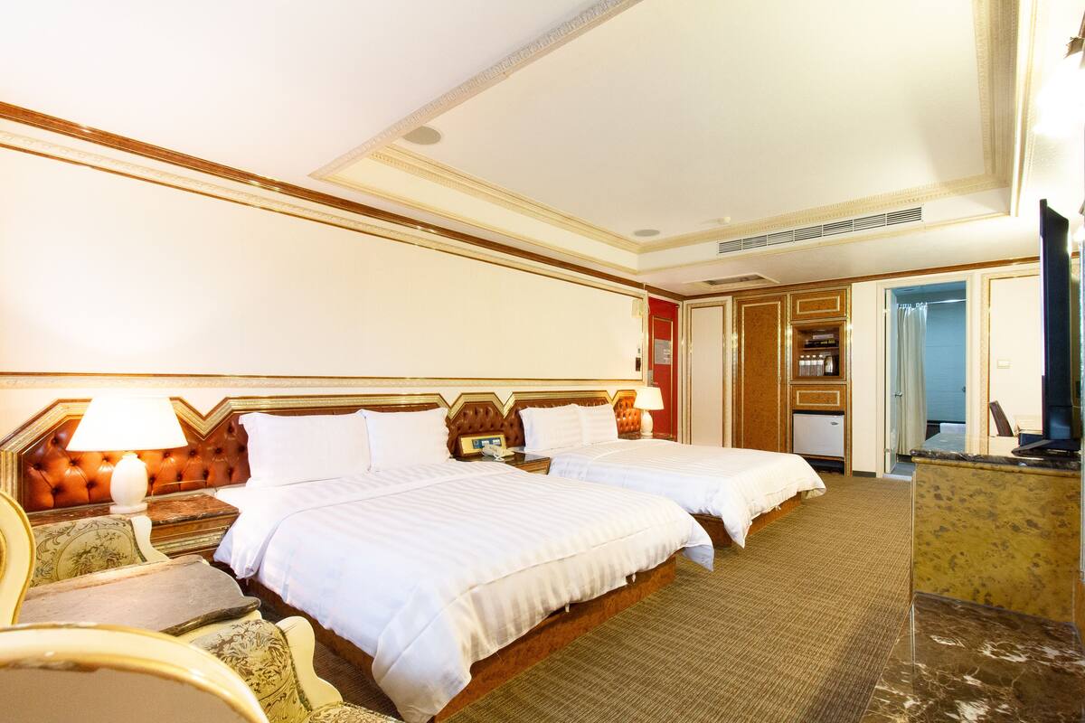 Habitación de hotel con dos camas, un escritorio, una silla, un televisor y un baño visible a través de una puerta abierta.