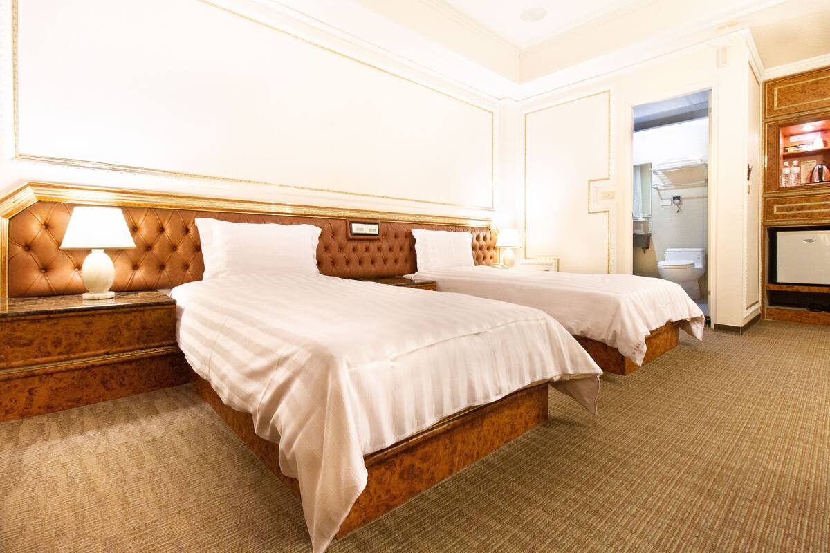 Habitación de hotel con dos camas, cabecera de madera, mesita de noche con lámpara y vistas al baño.