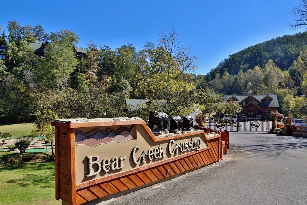 A wooden sign with "Bear Creek Crossing" written on it, surrounded by trees and a building in the background.