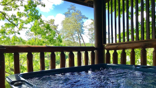 A hot tub with a wooden deck and railing, surrounded by lush greenery and trees.