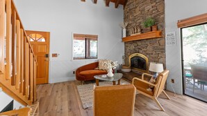 Un salon chaleureux avec une cheminée en pierre, des poutres apparentes en bois au plafond et un escalier en bois.