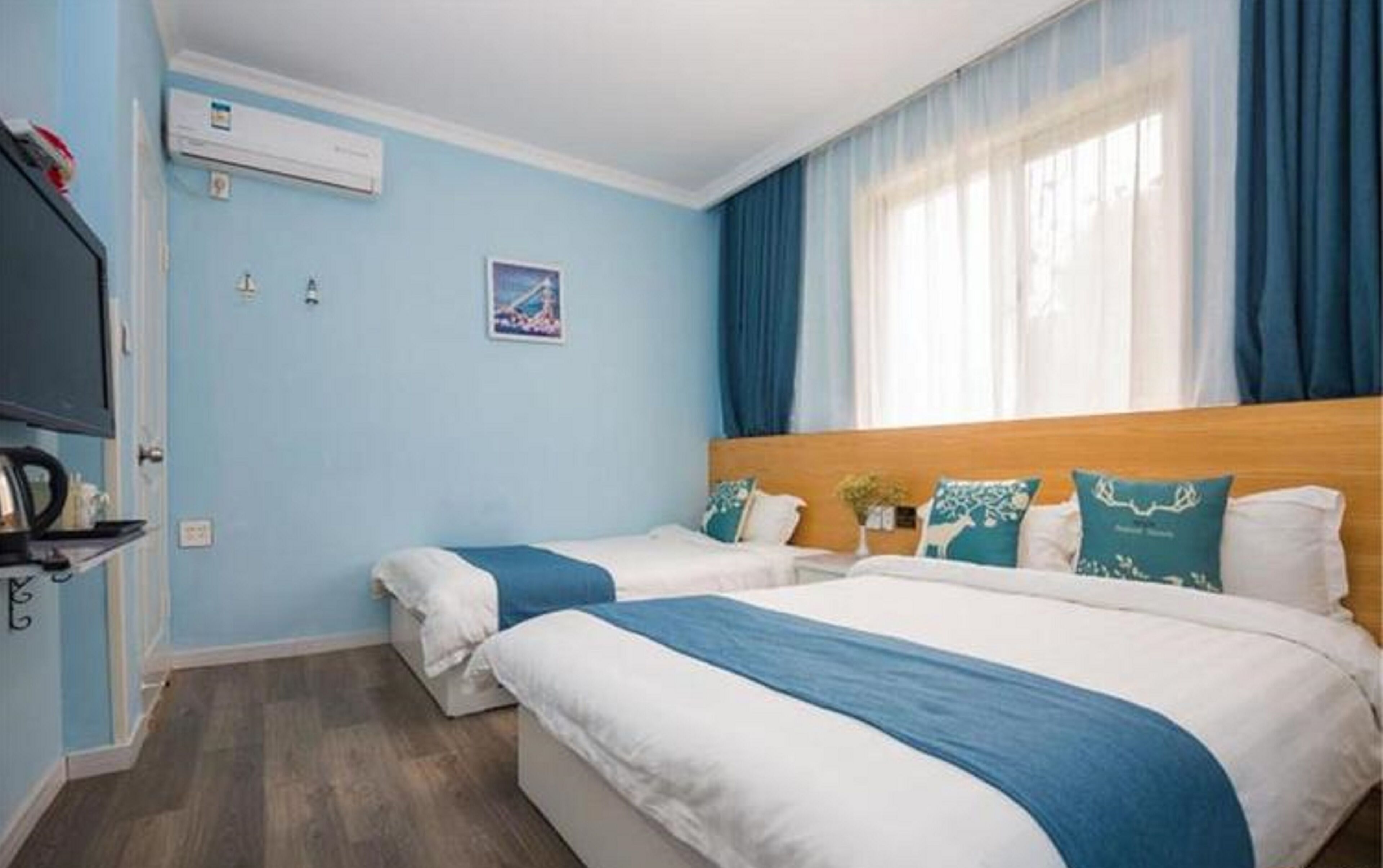 A hotel room with two beds, a flat-screen TV, an air conditioning unit, and a window with blue curtains.