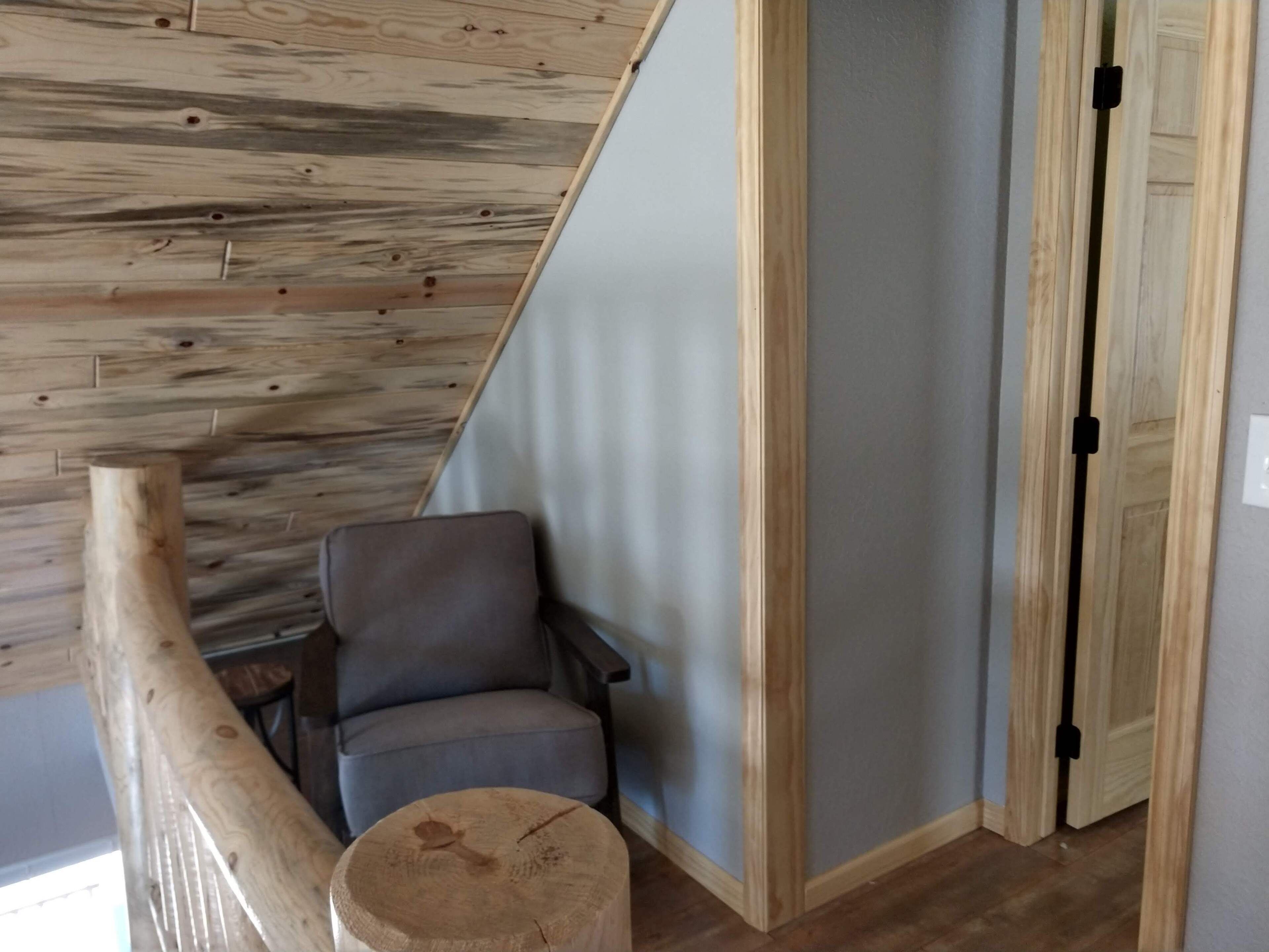 A room with a wooden ceiling, a grey chair, a wooden stool, and a door with a black handle.
