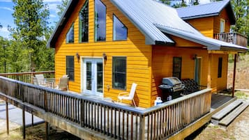 A two-story wooden cabin with a deck, outdoor seating, and a grill area.