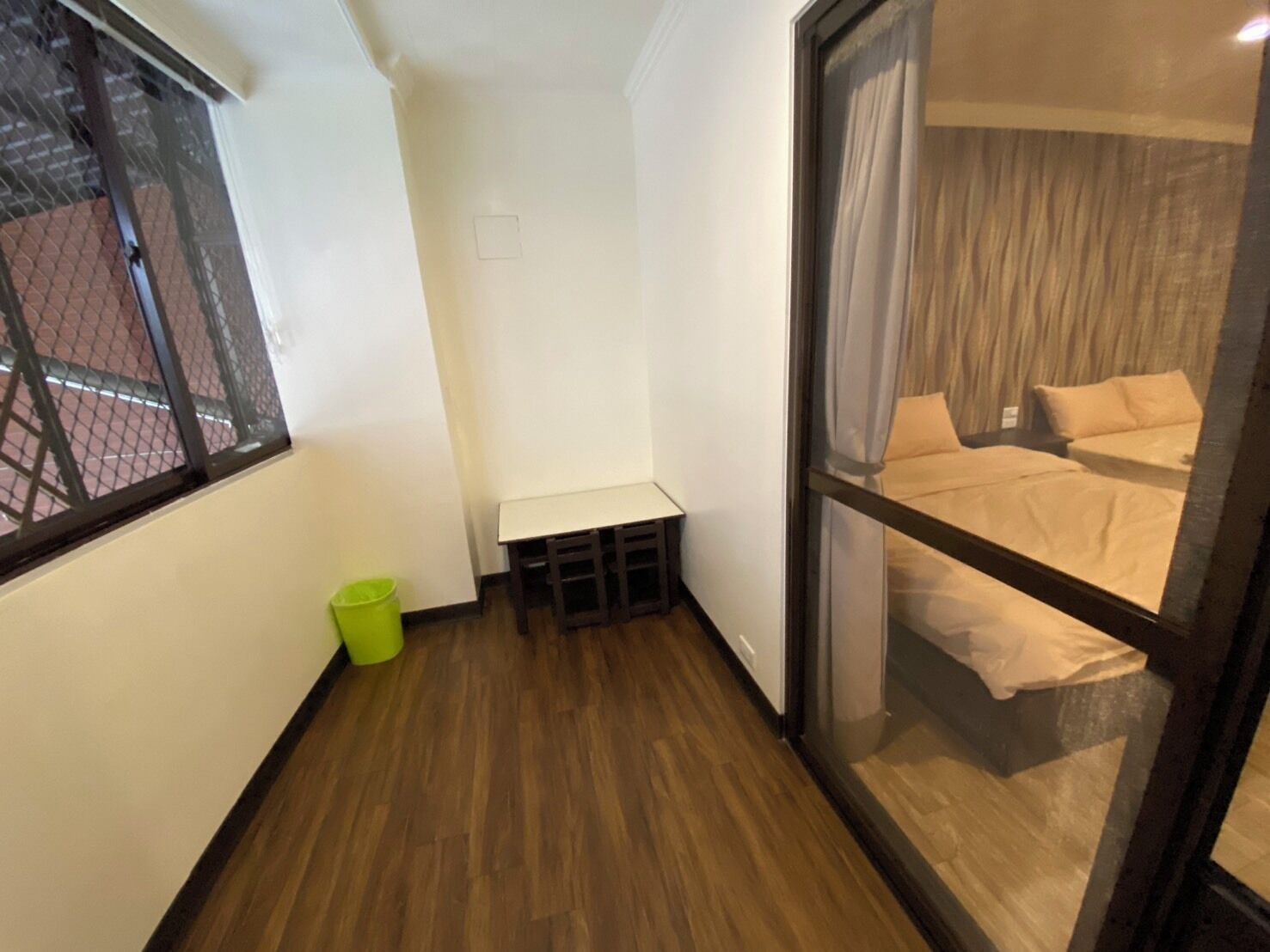 A small, narrow room with a wooden floor, a window with a metal screen, a small bench, and a trash bin.