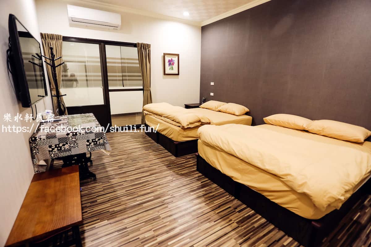 A hotel room with two beds, a wooden floor, a TV, a table, and a window with curtains.