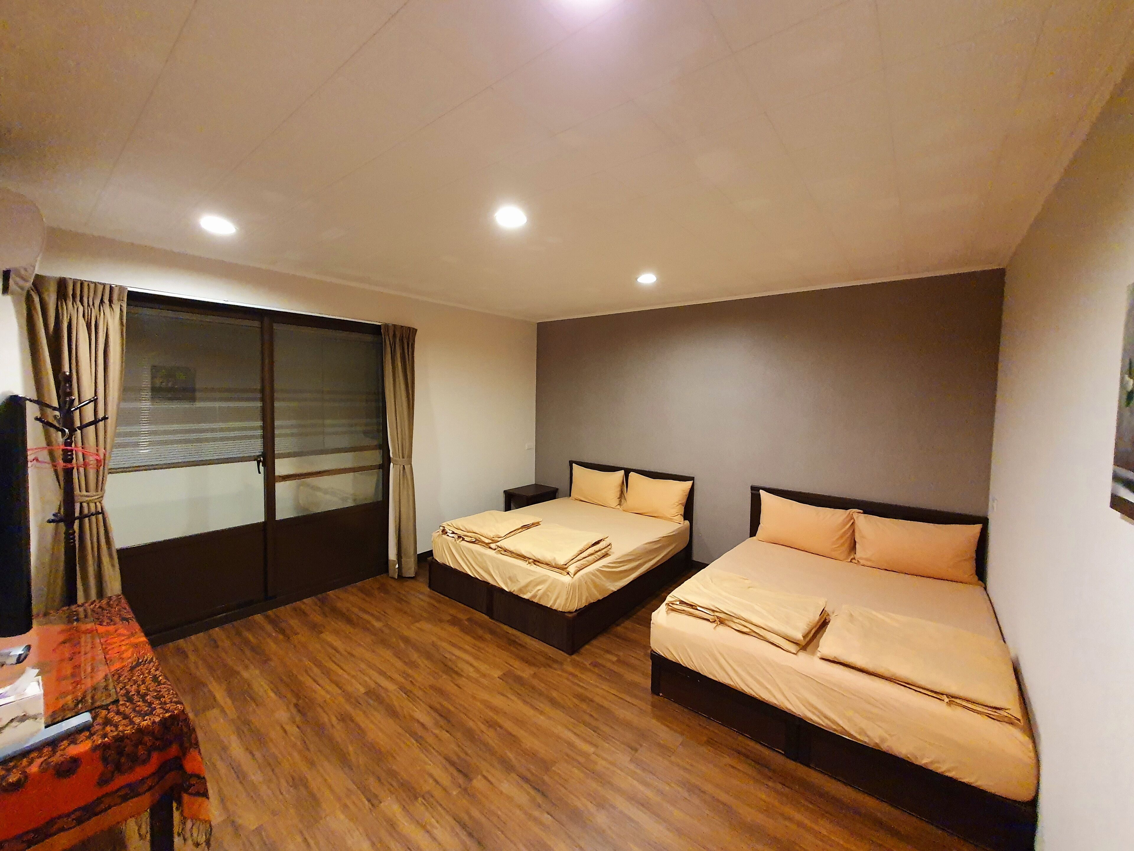 A hotel room with two beds, a wooden floor, a window with curtains, and a small table with a lamp.