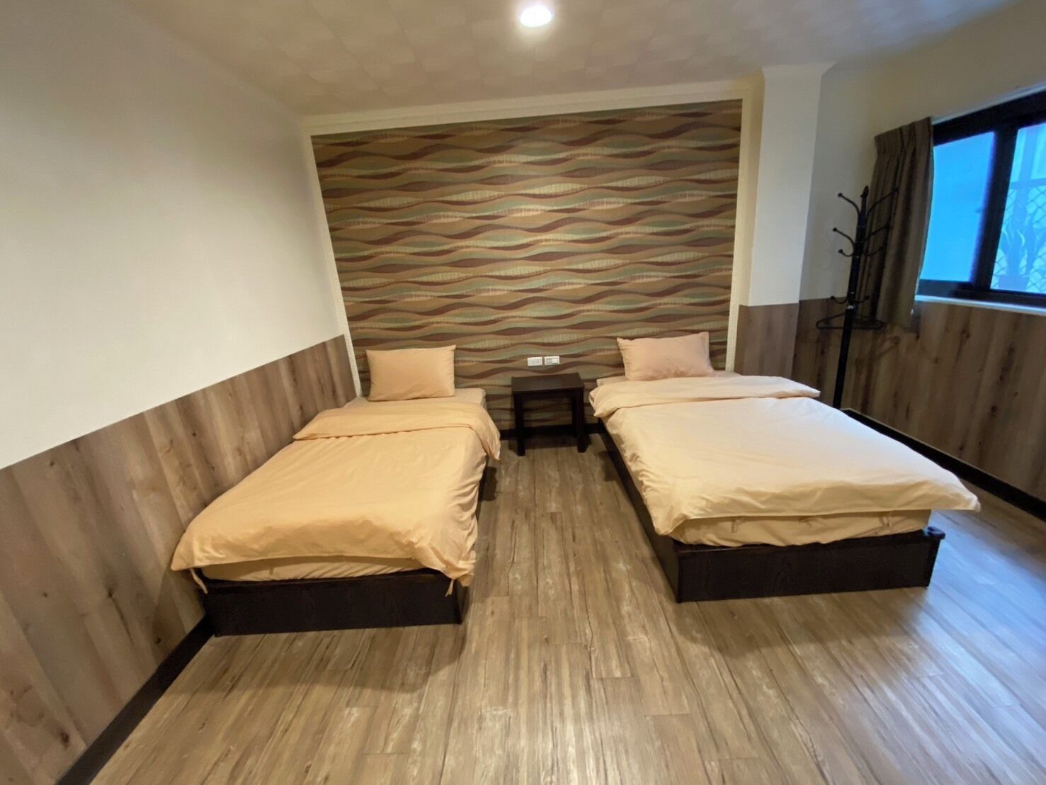 A room with two beds, a wooden floor, a patterned wall, and a window with curtains.