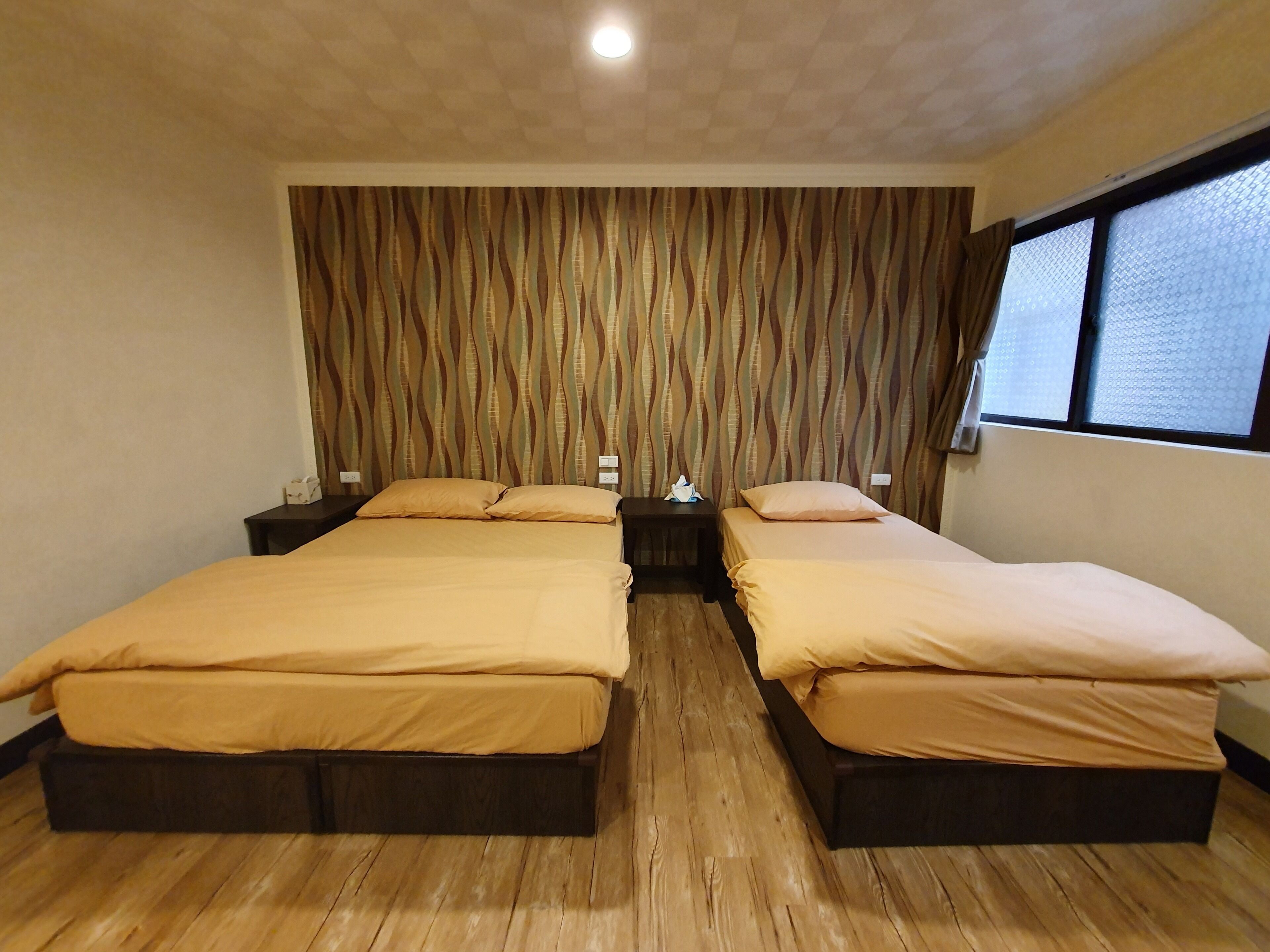 A double room with two beds, wooden flooring, a patterned curtain, and a window with blinds.