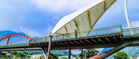 Un puente con un amplio toldo semejante a una vela, un elemento acuático con una esfera, y un telón de fondo montañoso.