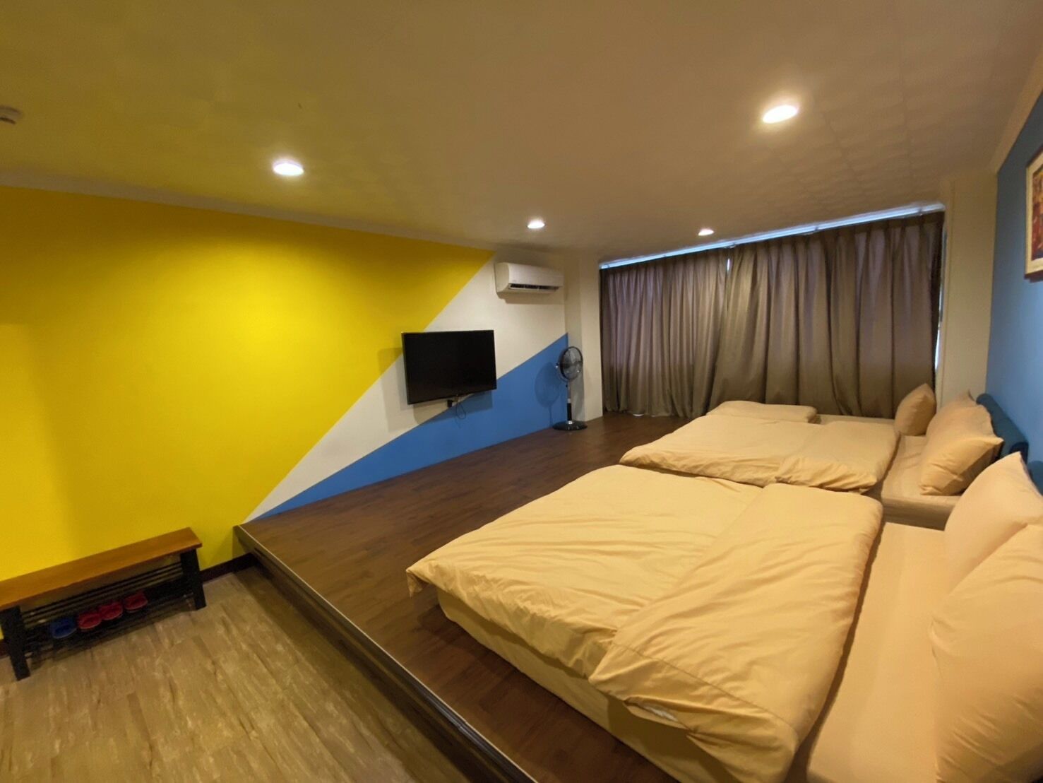 Habitación de hotel con una cama grande, un televisor de pantalla plana, un ventilador de pie y un banco con zapatos.