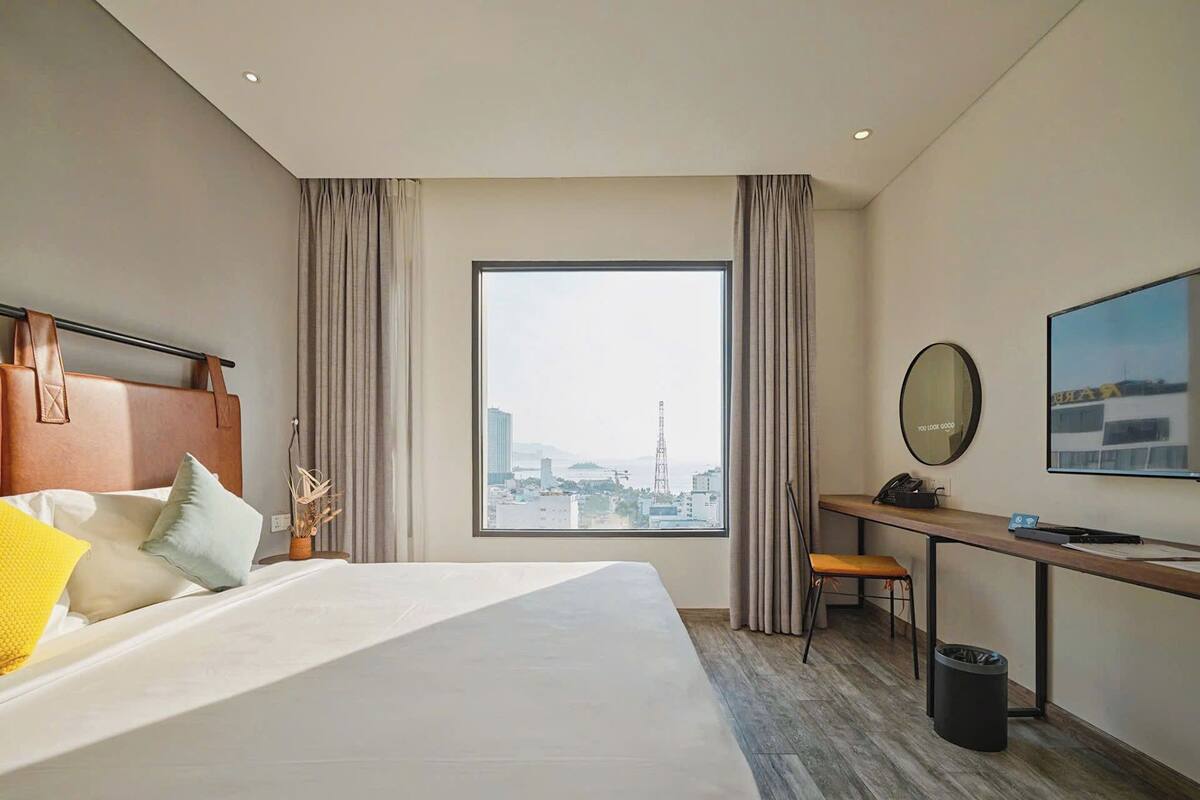 A hotel room with a large bed, a desk, a chair, a view of a cityscape, and a window with curtains.