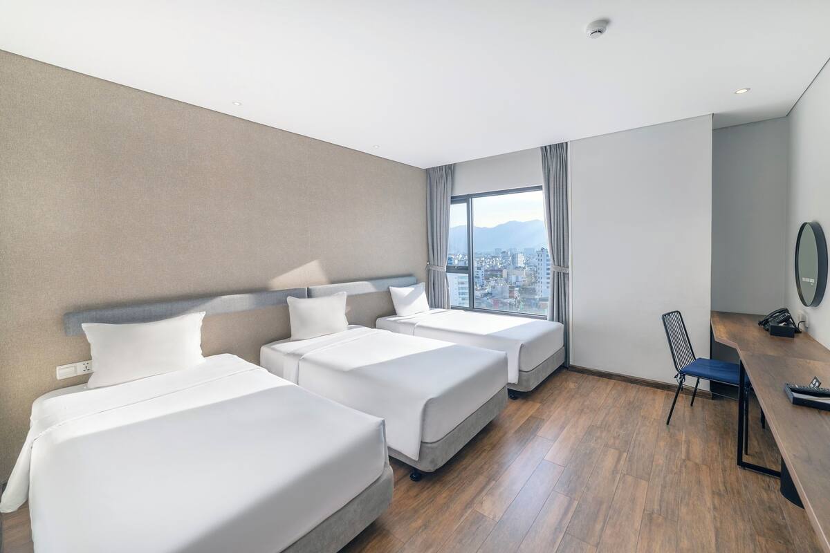 A hotel room with three beds, a desk, and a large window offering a city view.
