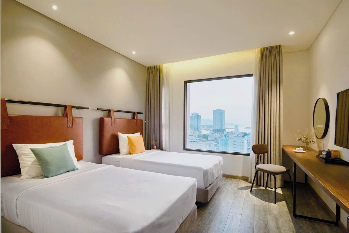 A hotel room with two beds, a desk, a chair, a large window with a city view, and a mirror on the wall.