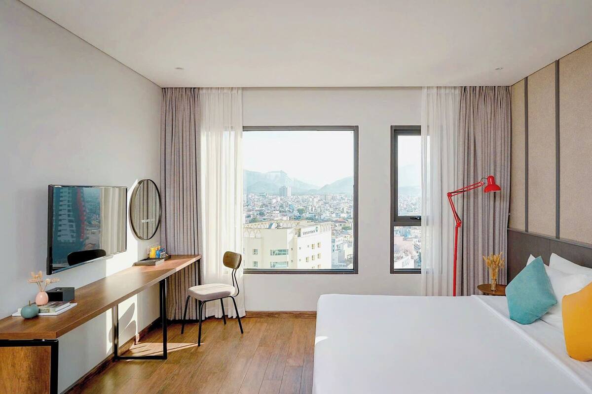 A hotel room with a bed, a desk, a chair, a TV, a red floor lamp, and a large window with a city view.