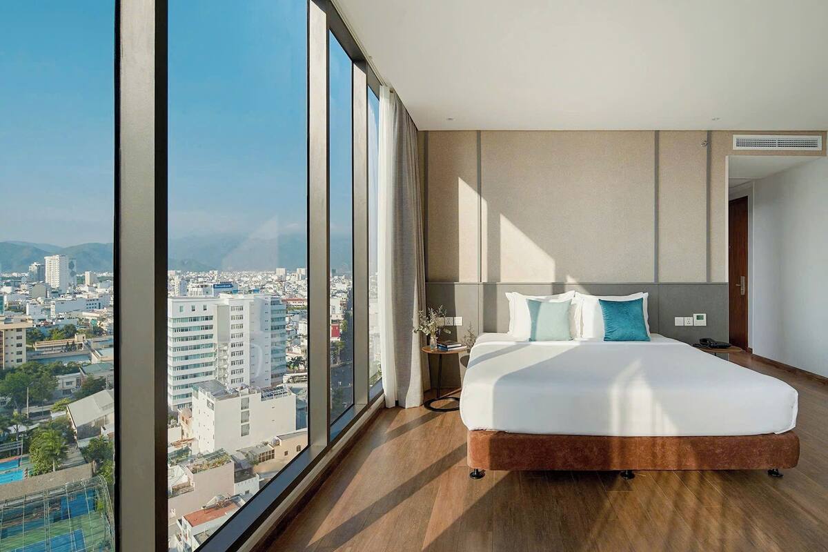 A modern hotel room with a large bed, a bedside table, and a view of a cityscape through floor-to-ceiling windows.