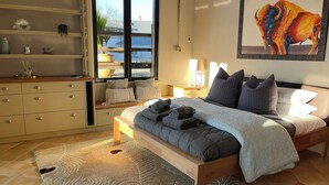 Un dormitorio con cama, mesitas de noche, un aparador, ventana con vistas, un cuadro y un estante empotrado con objetos decorativos.