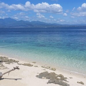 Una playa con agua turquesa cristalina, afloramientos rocosos y montañas distantes bajo un cielo parcialmente nublado.