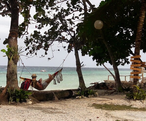 A hammock tied between trees on a beach with a signpost indicating directions to various locations. - Carolina del Mar (Samboan)