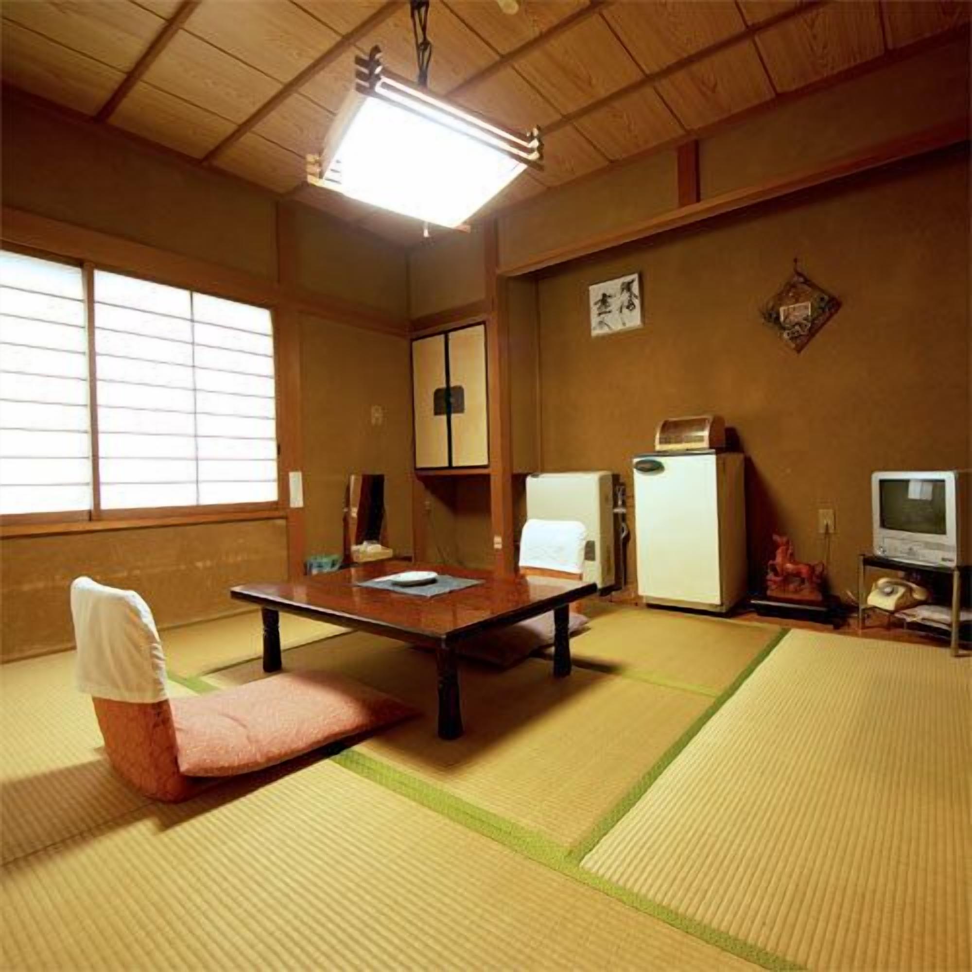 A traditional Japanese room with tatami flooring, a low table, a chair, a television, and a refrigerator.