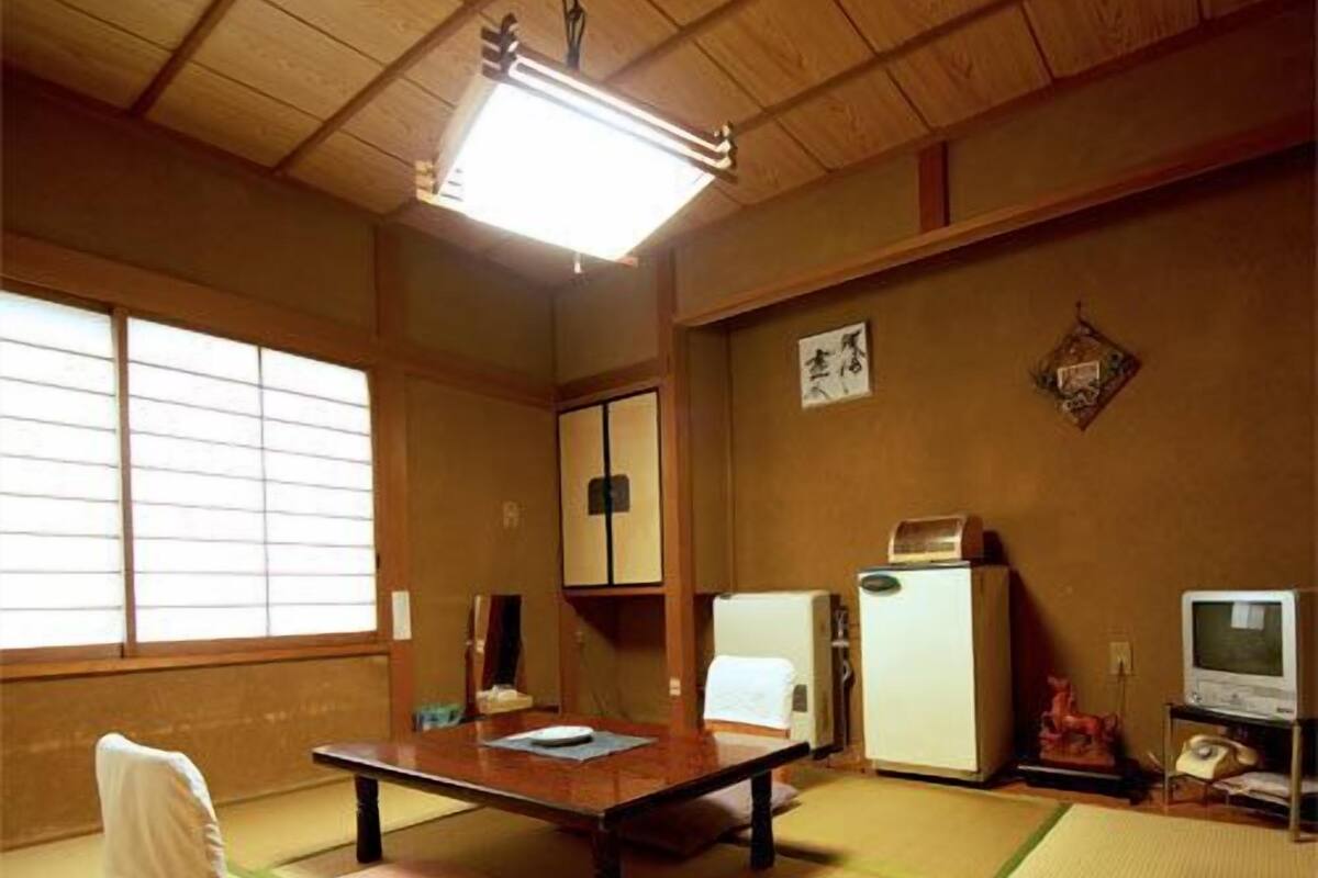 A traditional Japanese room with tatami flooring, a low table, a chair, a television, and a refrigerator.