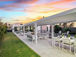 A covered outdoor patio with seating and tables, a swimming pool, and a modern building in the background.