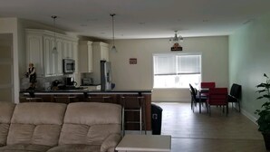 A spacious living room with a beige sofa, a small table, and a kitchen area with white cabinets and appliances. There is a dining area with red chairs and a window with blinds. - PRICE REDUCED Beautiful 3 Bedroom, 21/2 Bath Condo,walking distance to the beach (North Wildwood)