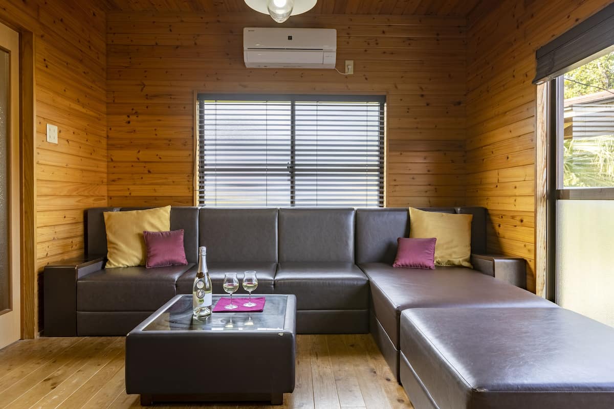 Ein Interieur einer Holzhütte mit einer Eckcouch, einem Glastisch und einem Fenster mit Jalousien.