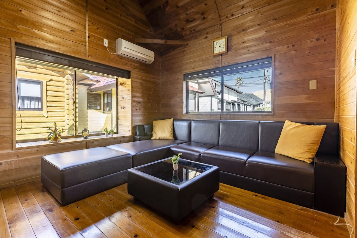 Ein Interieur einer Blockhütte mit einem Ecksofa, einem Glastisch, Holzdielen und großen Fenstern mit Blick auf ein Gebäude außerhalb.